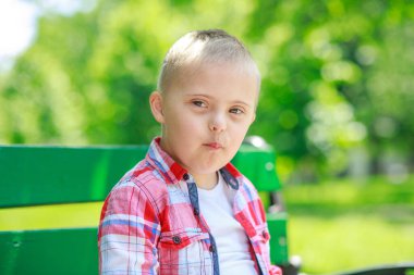 Parkta Down Sendromlu bir çocuk var. Yeşilliğin arka planına karşı. Çocuk gülümsüyor.