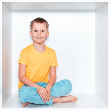 Sarı tişörtlü ve mavi pantolonlu şirin bir çocuk odasındaki mobilyalarda beyaz bir oyukta oturuyor. Kare beyaz bir kutuda bebek.