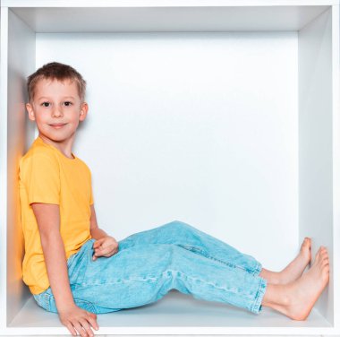 Sarı tişörtlü ve mavi pantolonlu şirin bir çocuk odasındaki mobilyalarda beyaz bir oyukta oturuyor. Kare beyaz bir kutuda bebek.