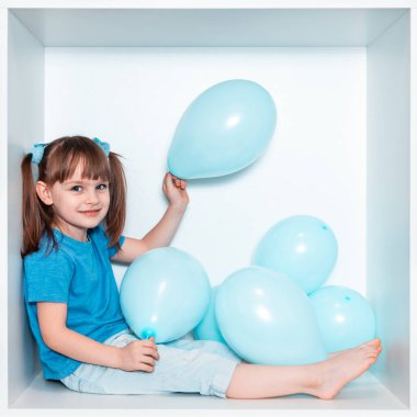 Mavi tişörtlü ve mavi pantolonlu şirin küçük bir kız odasında beyaz bir mobilya yuvasında oturuyor. Kare beyaz kutudaki bebek. Mavi balonlu kız.