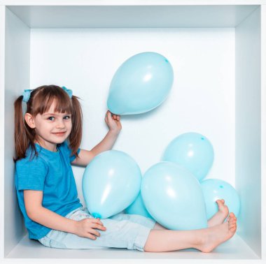 Mavi tişörtlü ve mavi pantolonlu şirin küçük bir kız odasında beyaz bir mobilya yuvasında oturuyor. Kare beyaz kutudaki bebek. Mavi balonlu kız.