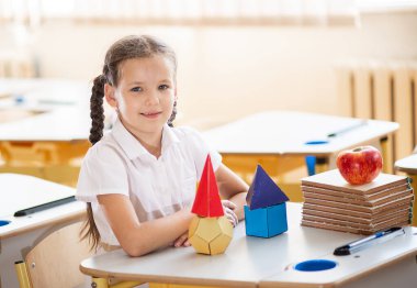 Okula geri dönmek, sınıftaki okul masasında oturan bir kız. Bir kız öğrenci geometrik şekilleri inceliyor - küp, piramit, top ve diğerleri. Okul malzemeleri bir çocuğun masasında..