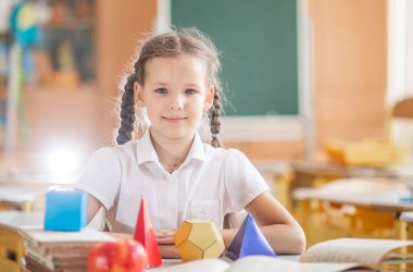 Okula geri dönmek, sınıftaki okul masasında oturan bir kız. Bir kız öğrenci geometrik şekilleri inceliyor - küp, piramit, top ve diğerleri. Okul malzemeleri bir çocuğun masasında..