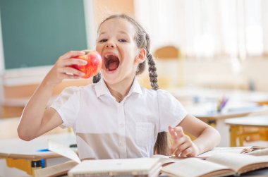 Okula geri dönersek, birinci sınıftan bir kız masasında oturmuş olgun bir elmayı ısırıyor. Ders sırasında atıştırmalık. Okuldaki çocuklar için uygun vitamin. Okul malzemeleri bir çocuğun masasında..