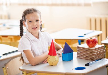 Okula geri dönmek, sınıftaki okul masasında oturan bir kız. Bir kız öğrenci geometrik şekilleri inceliyor - küp, piramit, top ve diğerleri. Okul malzemeleri bir çocuğun masasında..