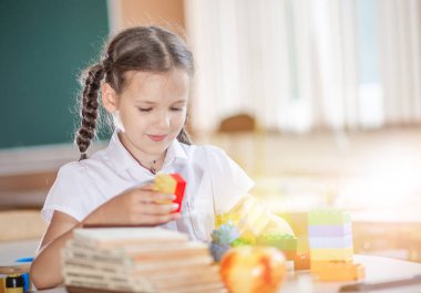 Okula geri dönmek, sınıftaki okul masasında oturan bir kız. Bir kız öğrenci geometrik şekilleri inceliyor - küp, piramit, top ve diğerleri. Okul malzemeleri bir çocuğun masasında..