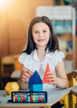 Okula geri dönmek, sınıftaki okul masasında oturan bir kız. Bir kız öğrenci geometrik şekilleri inceliyor - küp, piramit, top ve diğerleri. Okul malzemeleri bir çocuğun masasında..