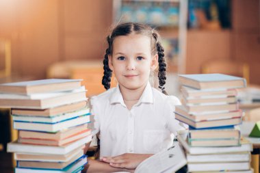 Bir kız masasında oturuyor ve iki uzun kitap yığını ve ders kitabı arasında neşeyle gülüyor. Küçük bir kız ödevini yapıyor. Okula dönelim. Sınav hazırlama konsepti.