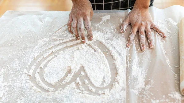 Hands of a young lady working with bakery flour and drawing heart shape on the table. Girl enjoys preparing materials for making bakery products