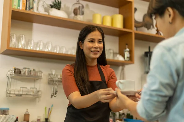 Young female coffee shop owner is holding a cup of hot coffee to customer with care. Lady with warm hospitality and care hands a cup of hot drink to her client in a small shop.