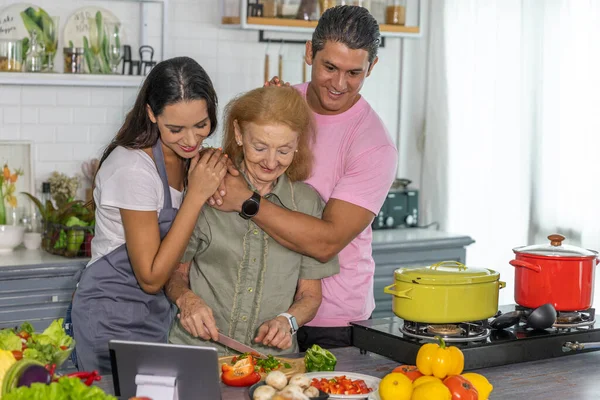 Young couple working with grand mom to take online cooking course and prepare meal at home. Happy family is cooking breakfast with healthy food together