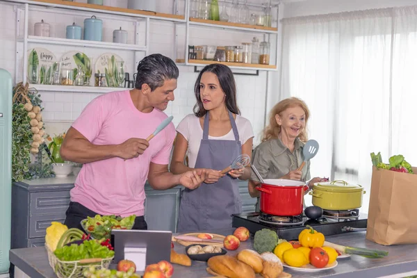 Young couple working with grand mom to take online cooking course and prepare meal at home. Happy family is cooking breakfast with healthy food together