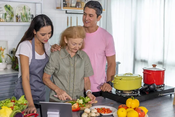 Young couple working with grand mom to take online cooking course and prepare meal at home. Happy family is cooking breakfast with healthy organic food together
