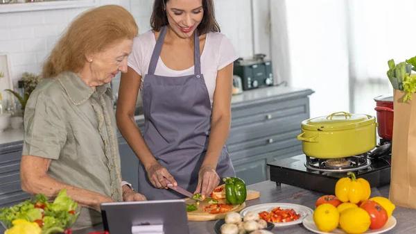 Elderly mother and young daughter getting online cooking class to prepare meal at home. Good family relationship between mom and daughter cooking together