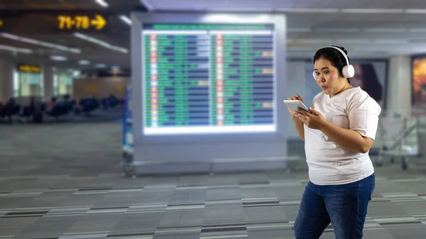 A young fat or overweight lady with headset enjoy listening to music and checking social media with mobile phone while waiting for her flight at the airport