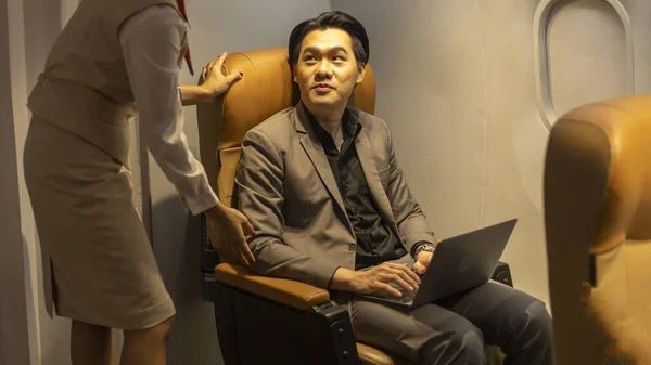 Young Asian male businessman gets service from a female flight attendant using laptop to connect with his colleagues while siting in an airplane during the flight