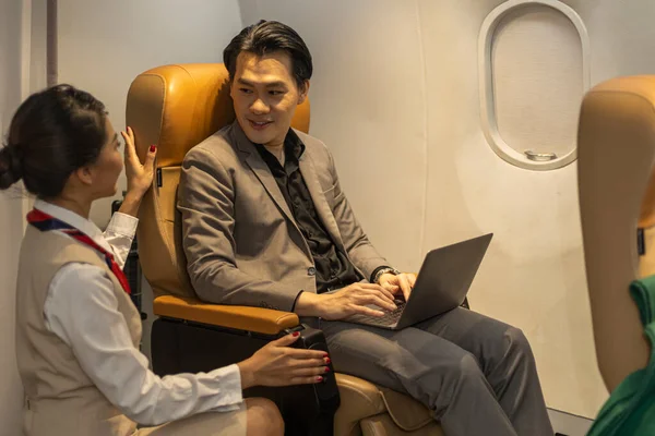 Young Asian male businessman gets service from a female flight attendant using laptop to connect with his colleagues while siting in an airplane during the flight
