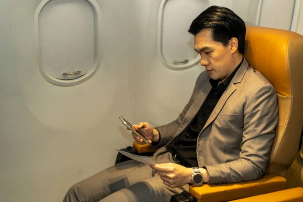 Young Asian male businessman uses mobile phone to connect with his colleague while siting in an airplane cabin before boarding
