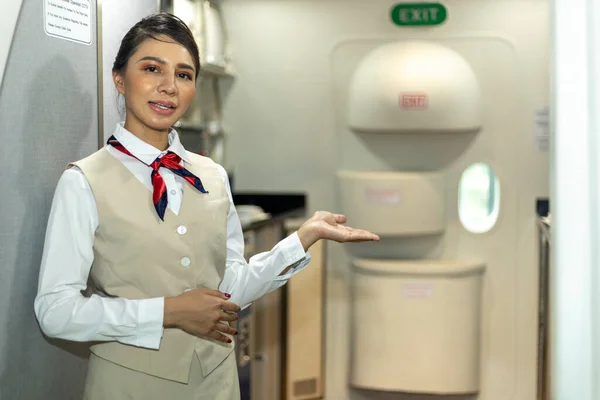 Young attractive airline attendant or stewardess is welcoming passengers to the flight at the airplane door. Hand gesture to ease customer or client