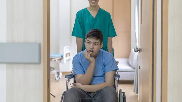 若い少年患者が車椅子に座って看護師のサポートを受ける 手術を受けた患者は 理学療法士のサポートを受けて車椅子を使用しています ストック写真