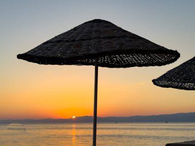 Güneş doğarken, ılık altın ışınları sakin okyanus sularını aydınlatır. Plaj şemsiyeleri, kumlu sahilde düzenli sıralar halinde durur. Resimli bir kıyı manzarası yaratırlar..