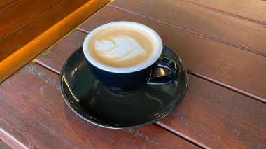 Siyah kapuçino bardağı, ahşap arka planda çay tabağı üzerinde latte sanatı. Bir fincan sıcak kapuçino ahşap masada. Latte üzerinde bir sanat eseri. Kahvaltı içeceği.