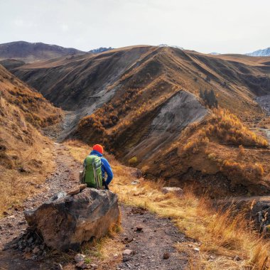 The dreaming hiker sits on a large boulder and happily looks into the distance against the background of blue dramatic clouds against the background of Caucasus mountains. Rest time on a hike. 