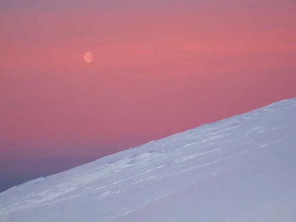 Atmospheric minimalist landscape with moon over high snowy mountain slope in dawn. Pink polar dawn sky with moon. Arctic evening sky.