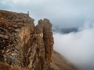 Soft focus. Tourist (man) stands on the cliff edge of Bermamyt plateau on high altitude under cloudy sky in foggy day. Rocks of Big Bermamyt - the so-called 