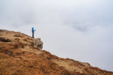 Tourist takes pics on mountain near abyss edge on high altitude under cloudy sky in foggy day. Man on high rock near precipice edge with wonderful misty view from above to large mountain range in away