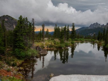 Gölde çarpıcı bir gün batımı. Bir yaz akşamı Ergaki 'deki Lazurnoye Gölü dramatik gökyüzü ve ağaç manzaralı Tayga kayaları arasında. Sibirya dağlarındaki Ergaki Doğa Parkı. 