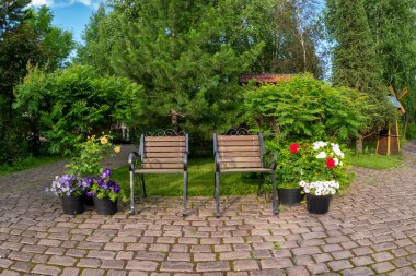 Bir yaz bahçesinde, taş döşeli kaide tasarımı konsepti üzerinde güzel çiçekli bitki taşlarıyla işlenmiş iki adet demirden koltuk..