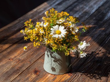 Parlak güneşin altında, ahşap bir masanın üzerinde beyaz papatyalar olan sarı kır çiçekleri buketi. Demir bir vazoda vahşi yaz çiçekleri..