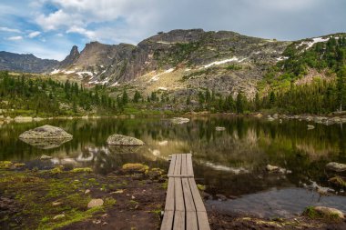 Tahta iskele suya iniyor. Ergaki 'deki Radughnoe Gölü' nde bir yaz taiga kayaları arasında mavi dramatik gökyüzü ve sedir ağaçları ile dağ manzarası. Sibirya 'daki Ergaki Doğa Parkı. 