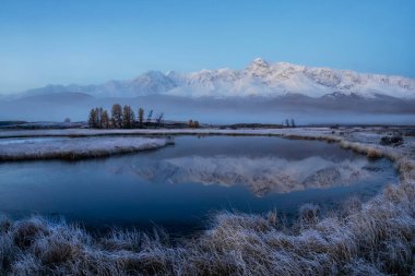 Dağlarda mavi saat. Kuru çimlerde sonbahar ayazı. Karlı tepeler bir dağ gölünün sakin soğuk suyuna yansıyor. Kuzey Chuysky Range ve Dzhangyskol Gölü şafakta. Rusya, Altai Cumhuriyeti.