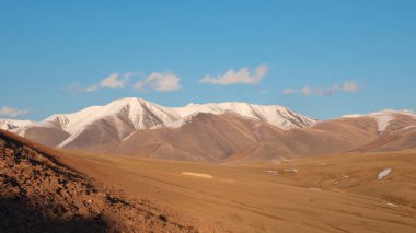 Parlak sonbahar dağ bozkırlarının geniş panoramik manzarası. Güneş ışığında geçitten dağ vadisine müthiş bir dağ manzarası ve ufukta mavi gökyüzünde beyaz bulutlar olan büyük dağ siluetleri..