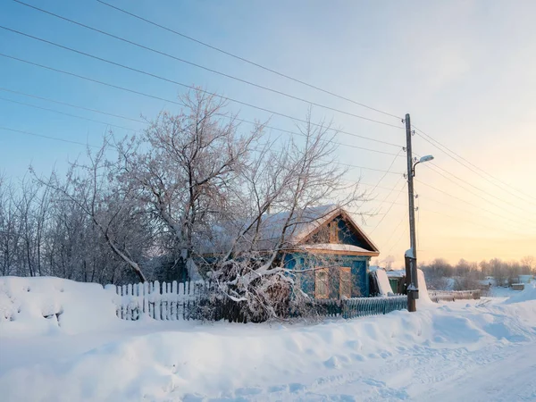 Rustic wooden house under the snow. Dawn on the outskirts of a Russian village in winter. Fence and an old electric pole in front of the house. Rustic landscape, real life. Perm Region.