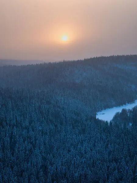 Sunrise over a snow-covered forest. Carpet of snowy coniferous forest under morning pink  frosty fog. Tops of hills are covered with frost. Haze to horizon hidden spruces. Vertical view.