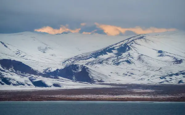 Snowy mountains above the turquoise waters of a large lake. View of the dark lake and snow hills under cloudy sky. Cold winter weather. Harsh climate background in Mongolia.