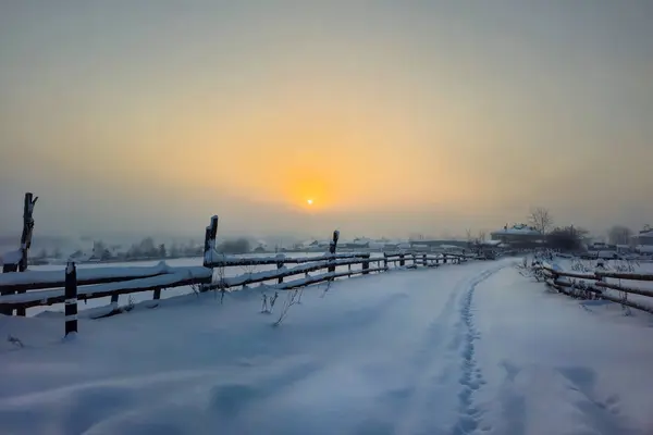 Beautiful winter morning. Frosty sunrise over the village. Misty sunrise over the field with wooden fence and village in the background. Snow path through snowdrifts.