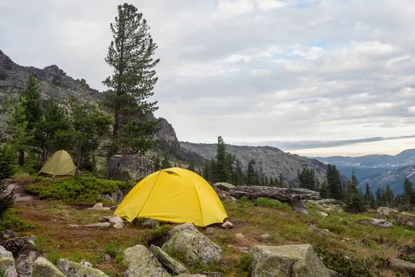 Dramatic landscape with two tents on forest hill among rocks and summer flora with view to large rocks range under cloudy sky. The tents and green summer colors in high mountains.