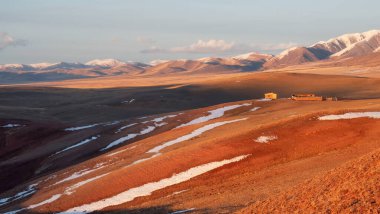 Parlak güneşli panoramik bozkır manzarası ve ahşap bir ahır. Kırmızı kuru otların üzerindeki ahşap ev, geniş bir çayır vadisinin dağ manzarası karla kaplı..
