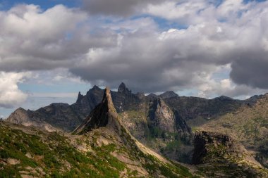 Alçak bulutların üstündeki sivri dağ zirveleriyle muhteşem dramatik bir manzara. Fırtınalı bulutlu gökyüzünde atmosferik geniş dağ tepeleri. Dev kayalar ve derin uçurumlar. Duvar için mükemmel yaz resmi.
