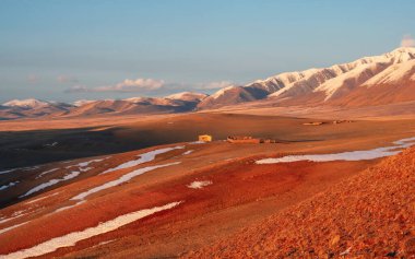 Kırmızı kuru çimenlerdeki ahşap ev, geniş bir çayır vadisinin dağ manzarası karla kaplı. Parlak güneşli panoramik bozkır manzarası ve ahşap bir ahır. Mükemmel sonbahar resmi.