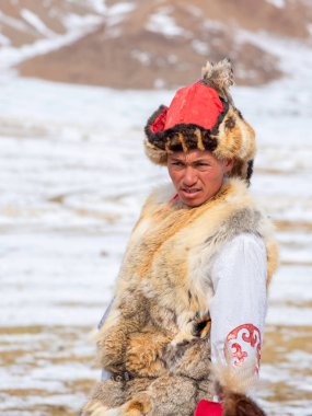 Ulgii, Moğolistan - 01 Ekim 2023: Kuzey Moğolistan. Moğolistan bozkırında bir Moğol erkek avcının portresi. Geleneksel giysileri olan genç Moğol bir kayanın yanında duruyor. Dikey görünüm.