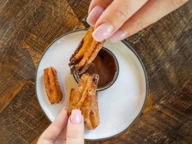 Kadın elleri hareket eder, şeker kaplı bir churro tutar, zengin bir çikolata sosuna batırır, hoşgörülü bir tatlı zevkini yakalar. Çikolata soslu tatlı Churros çöreği. Üst görünüm.