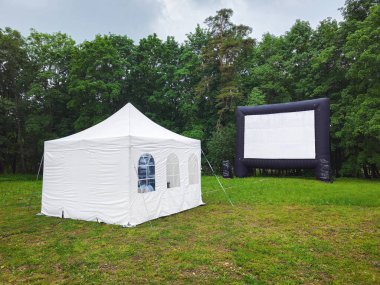 Beyaz küçük çadır ve parktaki çayırda büyük bir sinema perdesi. Halk parkındaki bir çayırda beyaz bir parti ya da etkinlik çadırı