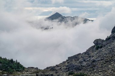 Keskin sarp kayalıklar mavi gökyüzündeki bulutların altındaki büyük dağlara karşı. Uzaktaki dev dağlarla çarpıcı katmanlı bir manzara. Muhteşem atmosferik sisli doğa manzarası..