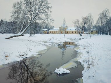 Kış köyü. Karlı genişler. Nehir. Kış manzarası. Sudaki yansımalar. Karla kaplı parktaki ağaçların arasından küçük nehir akar. Gatchina 'daki Eski Çiftlik Binası. 