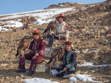 Ulgii, Moğolistan - 30 Eylül 2023: Kayalık güneşli bir dağ yamacında altın kartallı üç mutlu avcı poz veriyor. Kuzey Moğolistan.